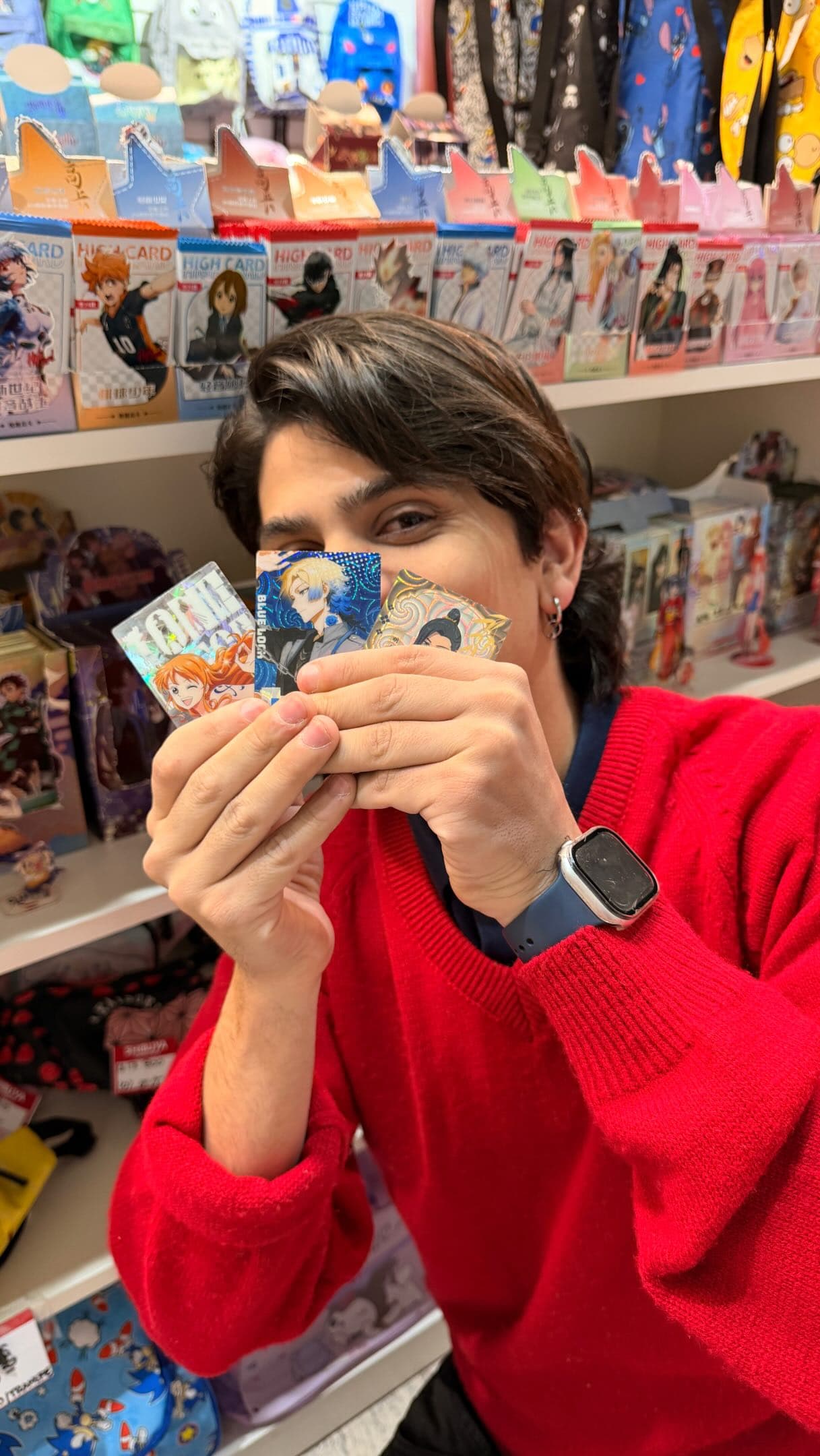 Foto de Shibuya: una persona con buzo rojo sostiene un abanico de cartas frente a la cara, con las estanterías de figuras y cajas del local detrás.