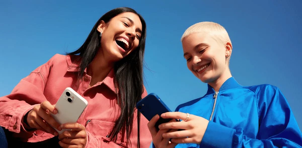 Dos amigas se ríen mirando un teléfono sobre un fondo azul