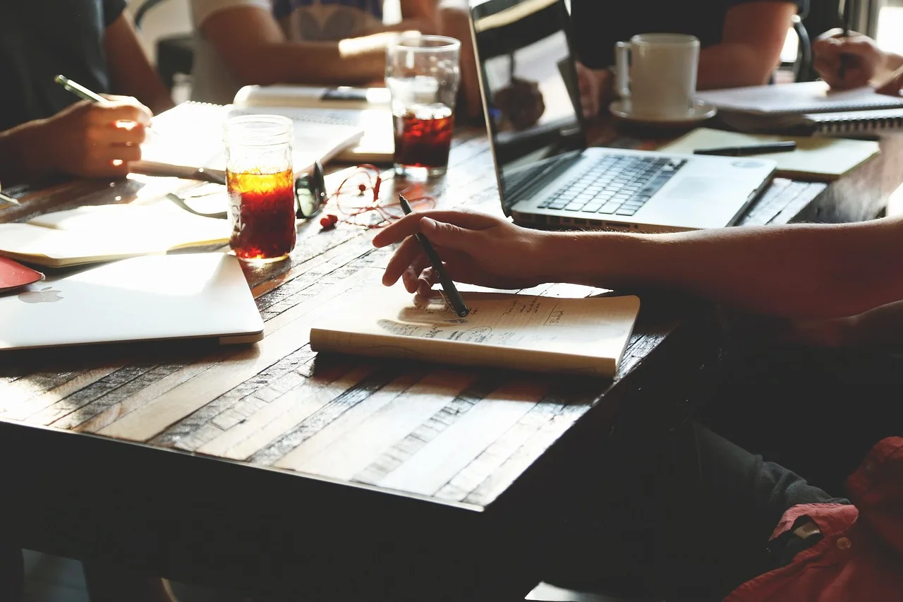 Varias personas trabajan alrededor de una mesa de madera con computadoras portátiles, cuadernos y vasos