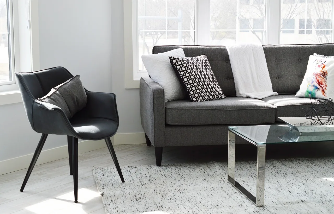 Un sillón gris con almohadones y una mesa ratona de vidrio en un living iluminado por ventanales