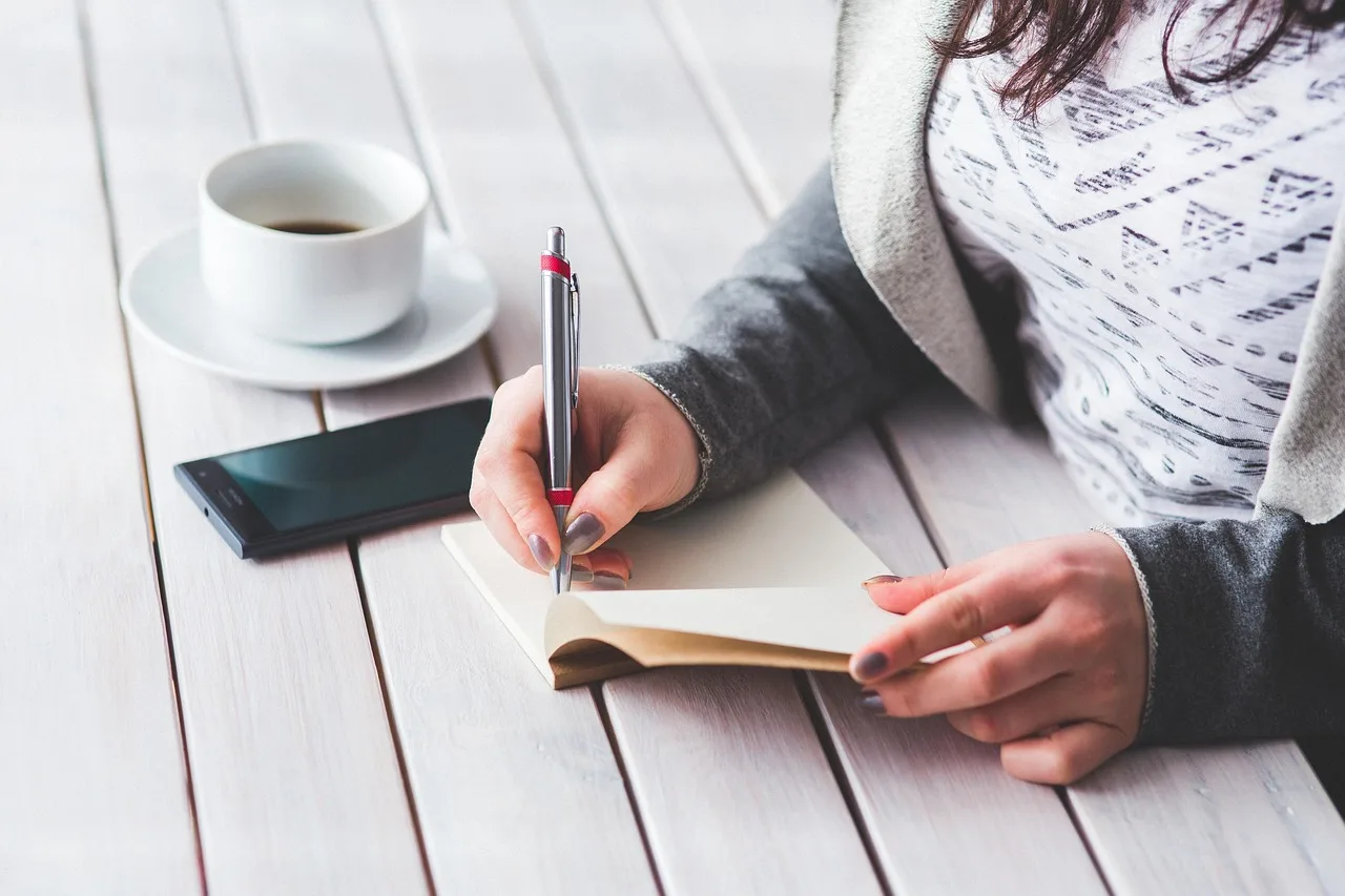 Una persona anota en una libreta sobre una mesa de madera blanca, con un teléfono y una taza de café al lado