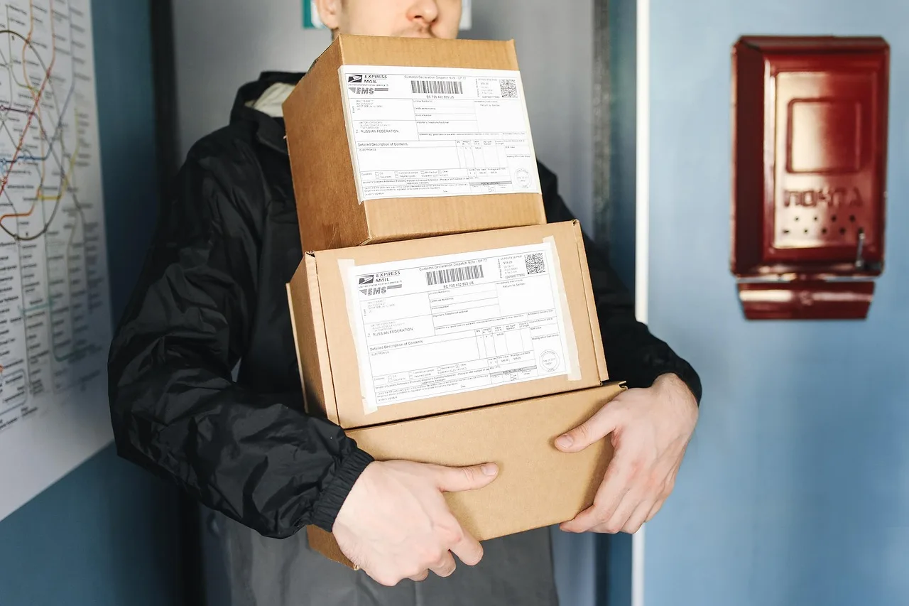 Una persona sostiene tres cajas de cartón apiladas con etiquetas de envío junto a la puerta de entrada