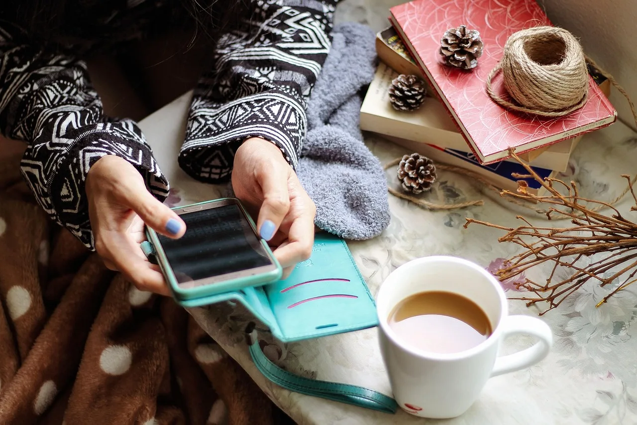 Unas manos sostienen un teléfono sobre una manta junto a una taza de café y unos libros