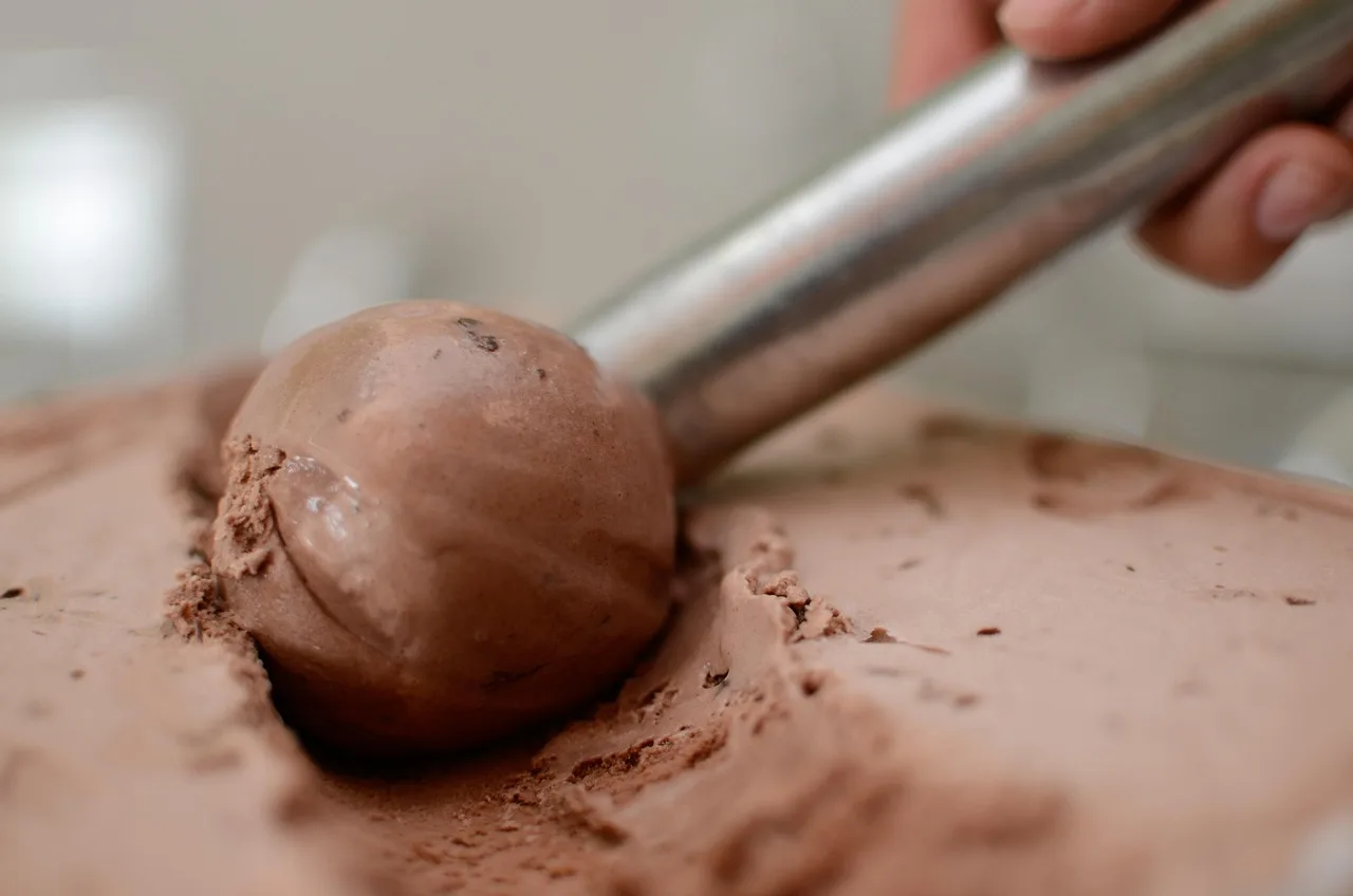 Una cuchara de metal saca una bocha de helado de chocolate de una bandeja recién empezada