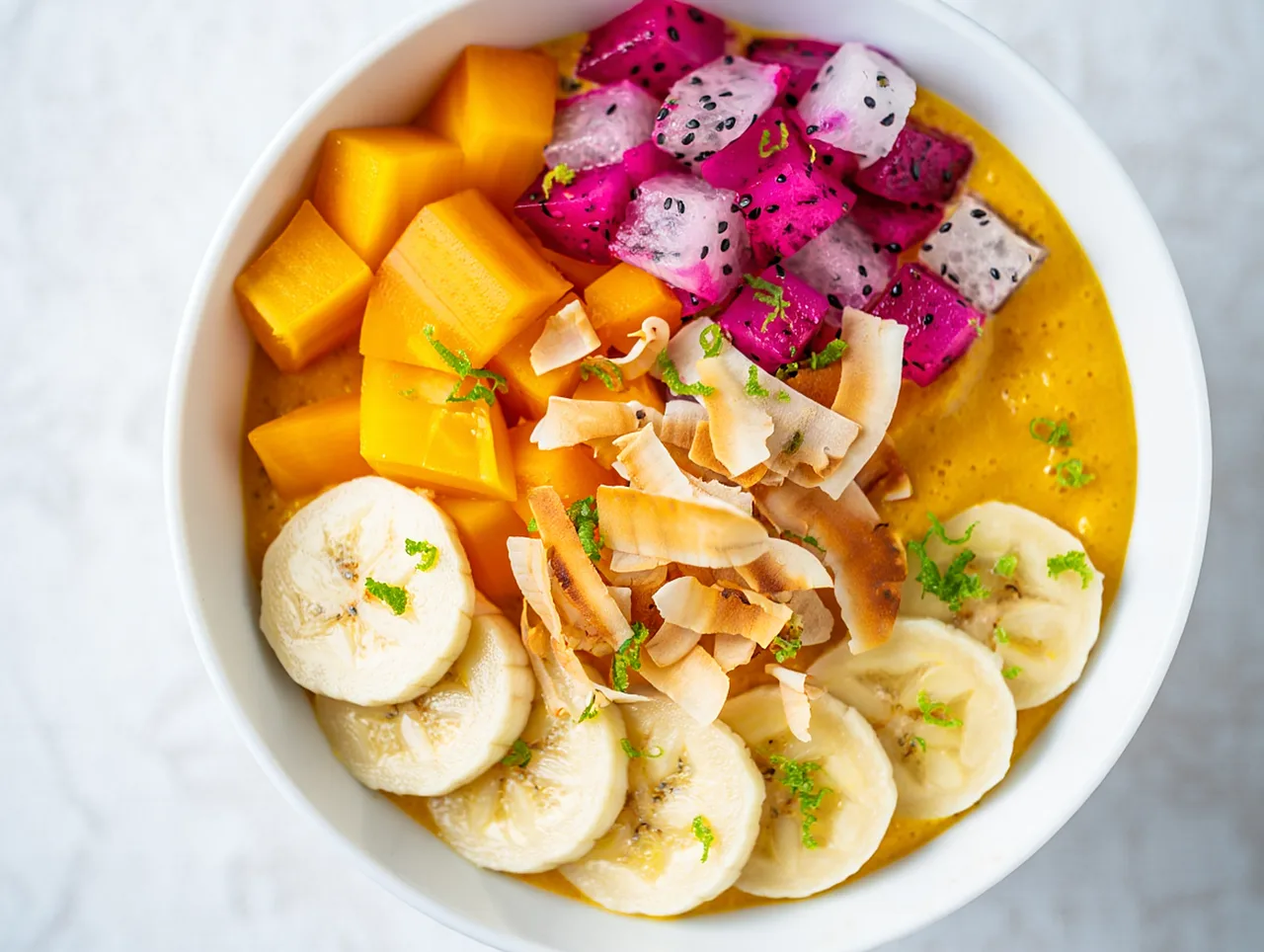 Un bowl de licuado de mango con banana, pitaya y coco en láminas visto desde arriba sobre una mesa clara