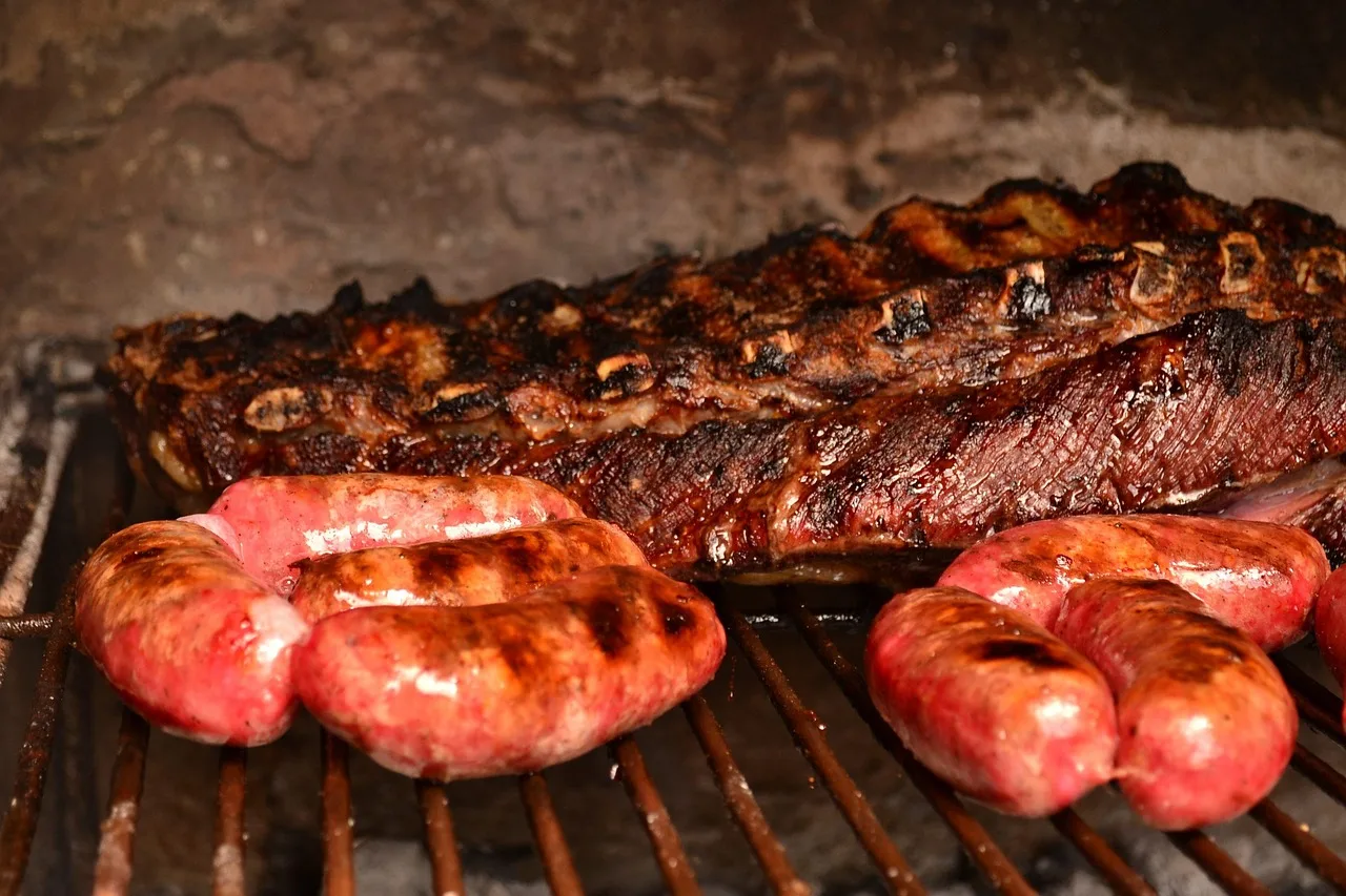 Una tira de asado y varios chorizos dorándose sobre una parrilla de hierro frente a una pared de piedra