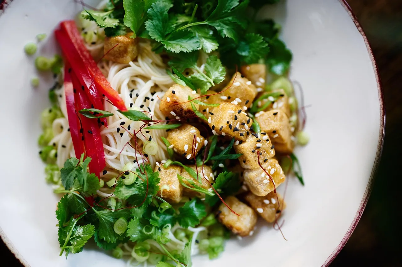 Un plato hondo con fideos, cubos de tofu dorado, hojas de cilantro y tiras de morrón rojo