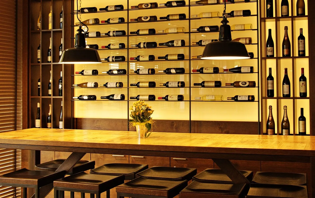 Una mesa larga de madera con banquetas frente a una pared iluminada llena de botellas de vino acostadas