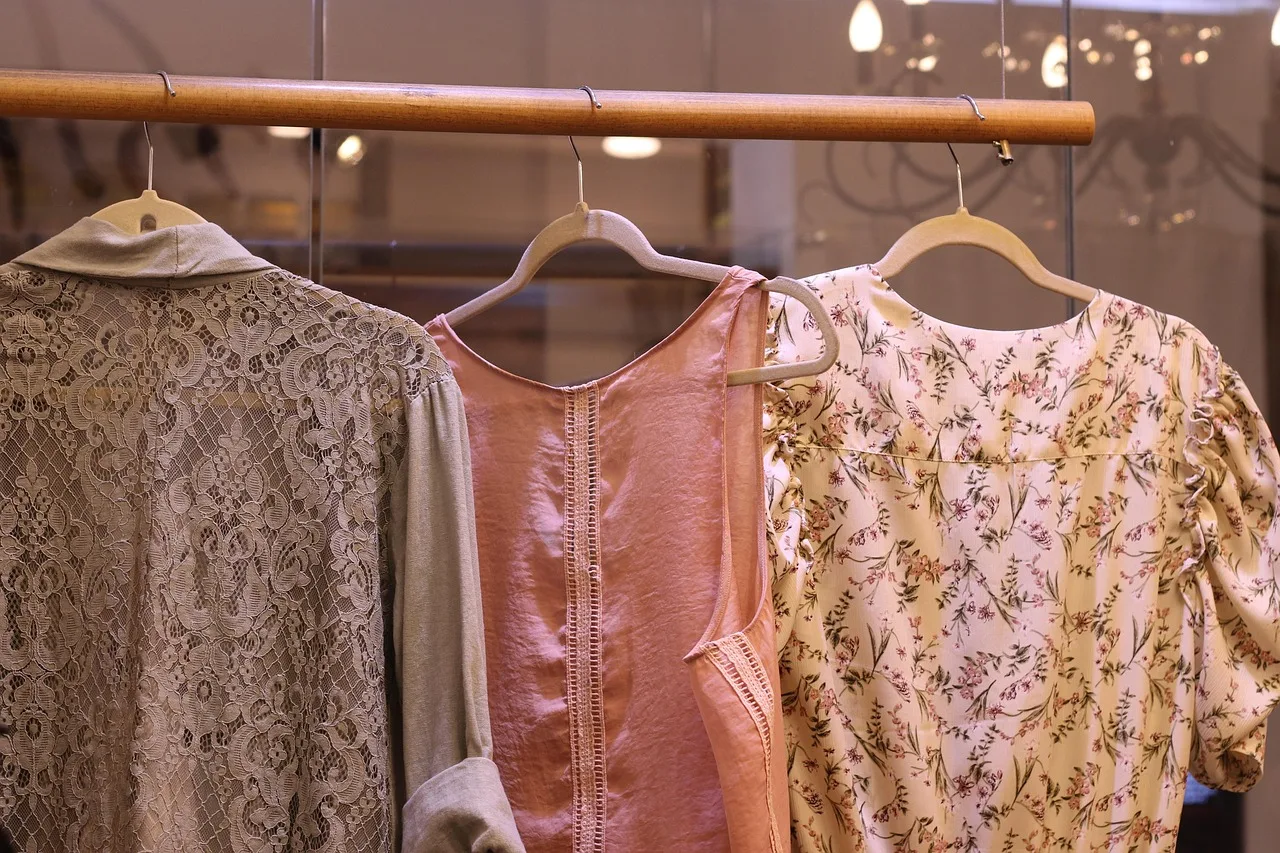 Tres blusas de telas claras cuelgan de perchas en una barra de madera dentro de una tienda de ropa